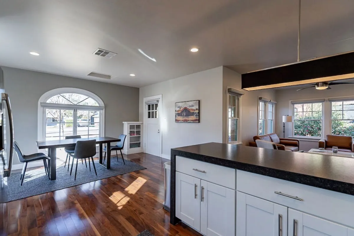 This spacious interior shot showcases a welcoming dining area with a large table positioned near a wide window, adjacent to a comfortable sofa lounge. The room is bathed in natural light, emphasizing warm wood tones and a relaxed, communal vibe ideal for shared meals or planning adventures.
