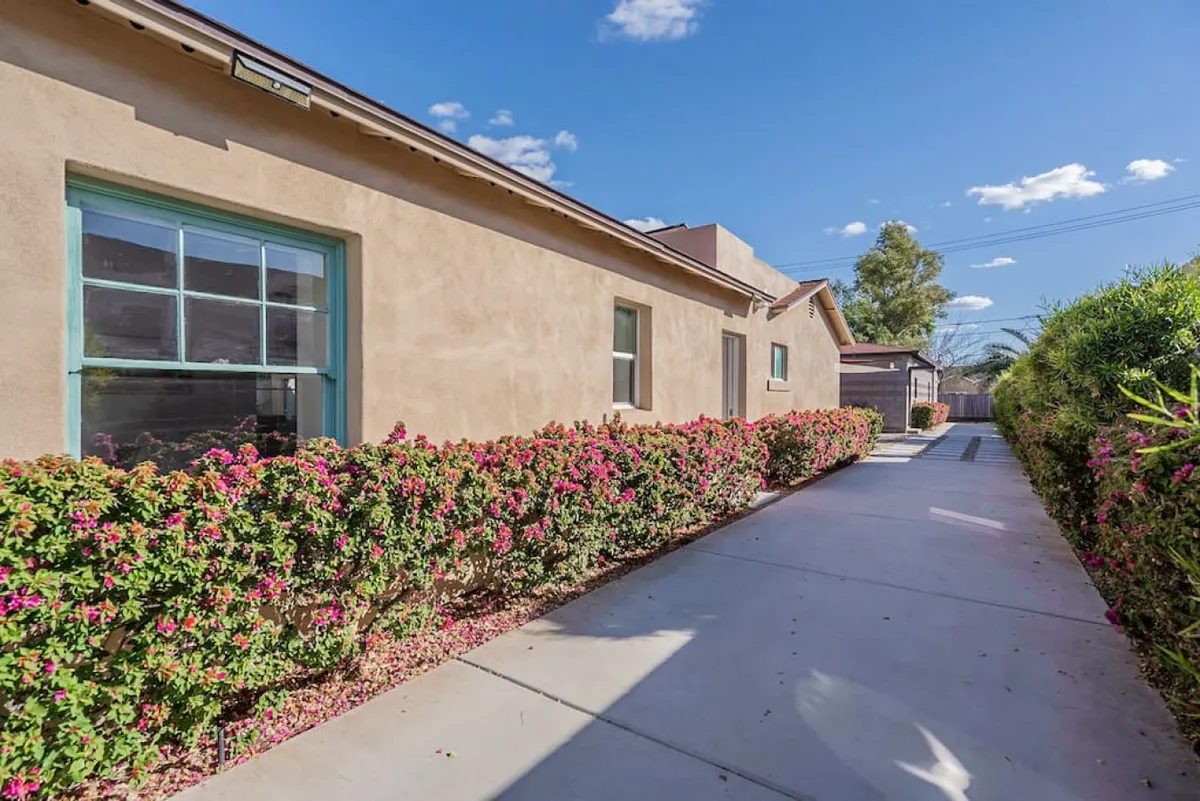 The image presents a charming exterior pathway lined with flowering bushes leading to the building’s facade. The landscaping and patio area create a cheerful, welcoming ambiance for arrivals and morning strolls.