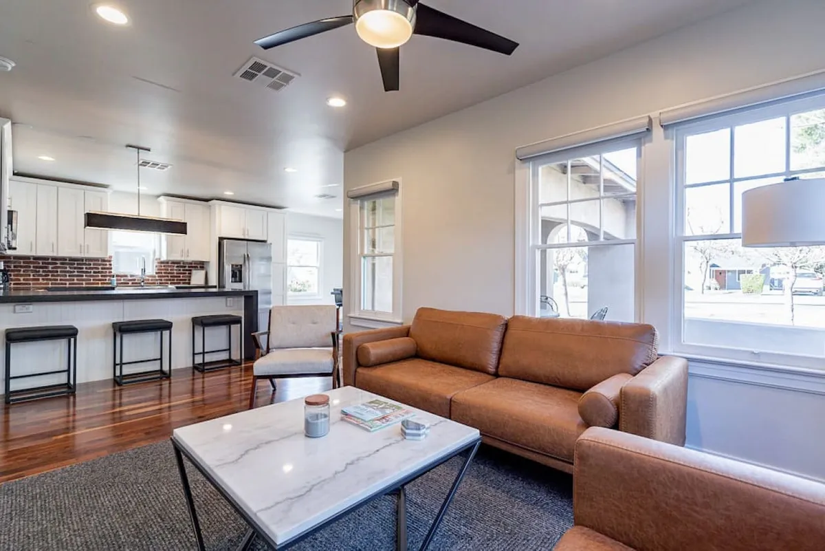 A welcoming living area featuring a comfortable sofa, a coffee table perfect for gathering around, a kitchen bar in view, and large windows that let natural light flood the space. The decor suggests a place ideal for socializing, relaxing with friends, and planning outings.