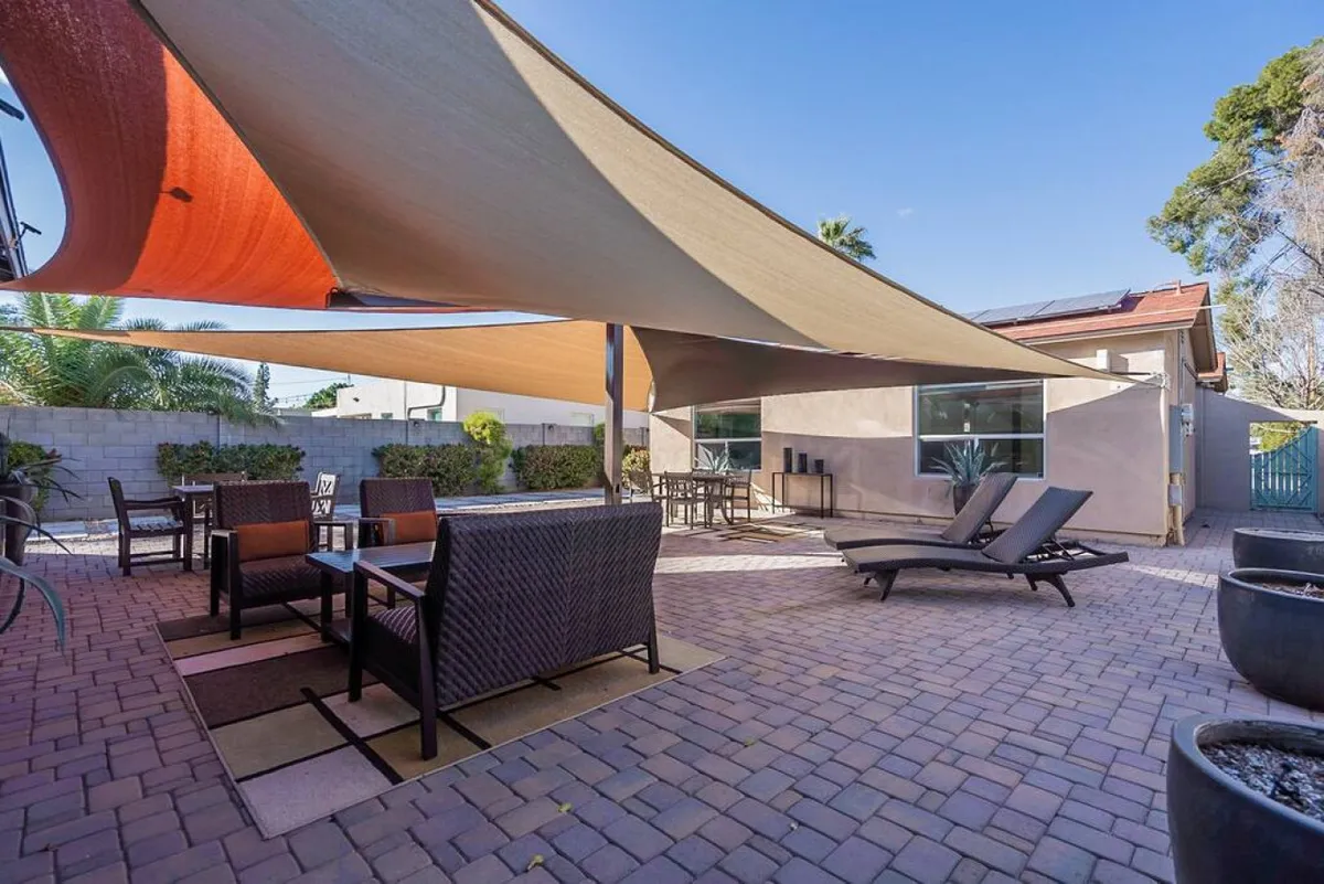 A spacious shared courtyard with a dining area, multiple lounging chairs, and shade sails overhead. The landscaped border and greenery create an inviting, social atmosphere ideal for group gatherings, outdoor meals, or relaxation under the Arizona sky.