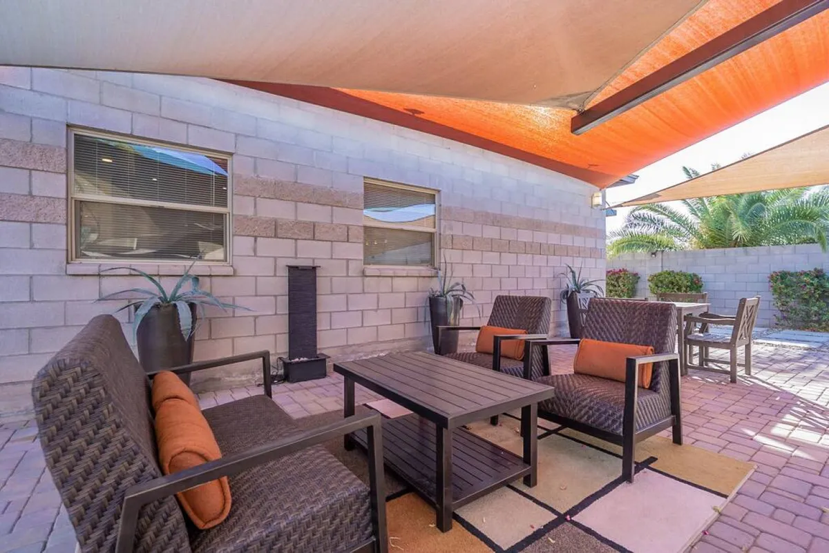 A welcoming outdoor seating area features a coffee table, comfortable chairs, and shade sails above, surrounded by lush potted plants. The space is arranged for conversation, relaxation, or enjoying drinks outdoors.