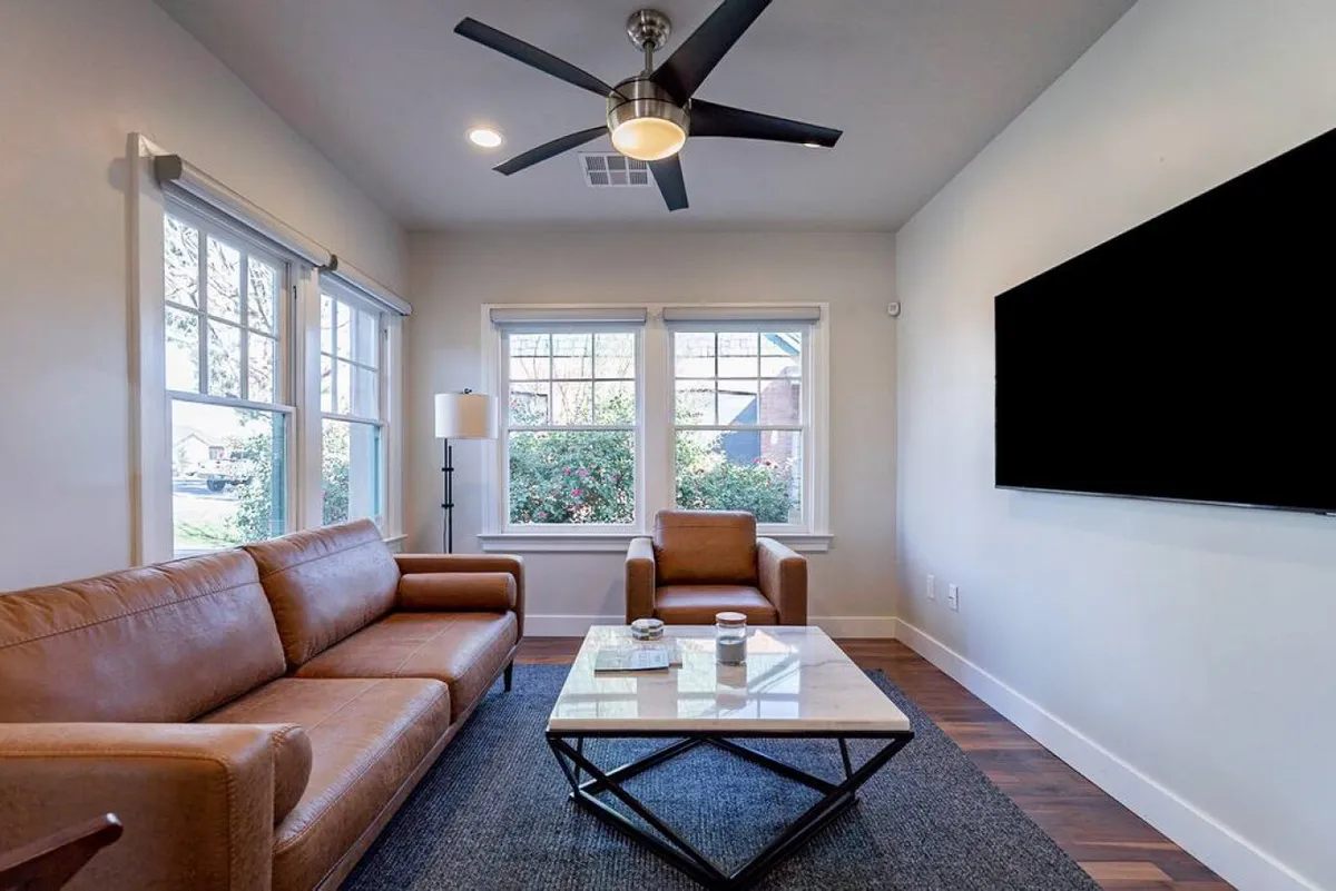 A cozy seating area with a brown leather sofa and large windows that let in plenty of light. The scene exudes comfort, with casual seating for conversation or watching TV, and direct views to the outside, suggesting indoor-outdoor flow.
