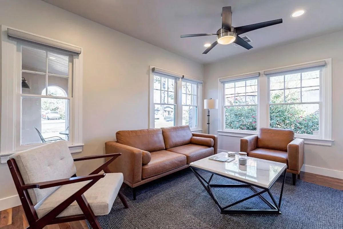 This inviting interior image features a leather sofa and glass coffee table, with large windows allowing abundant sunlight to fill the space. The setting is both contemporary and comfortable, with original architectural touches evident in the window design and ceiling fan, while plush furnishings create a cozy atmosphere for relaxing or conversation.