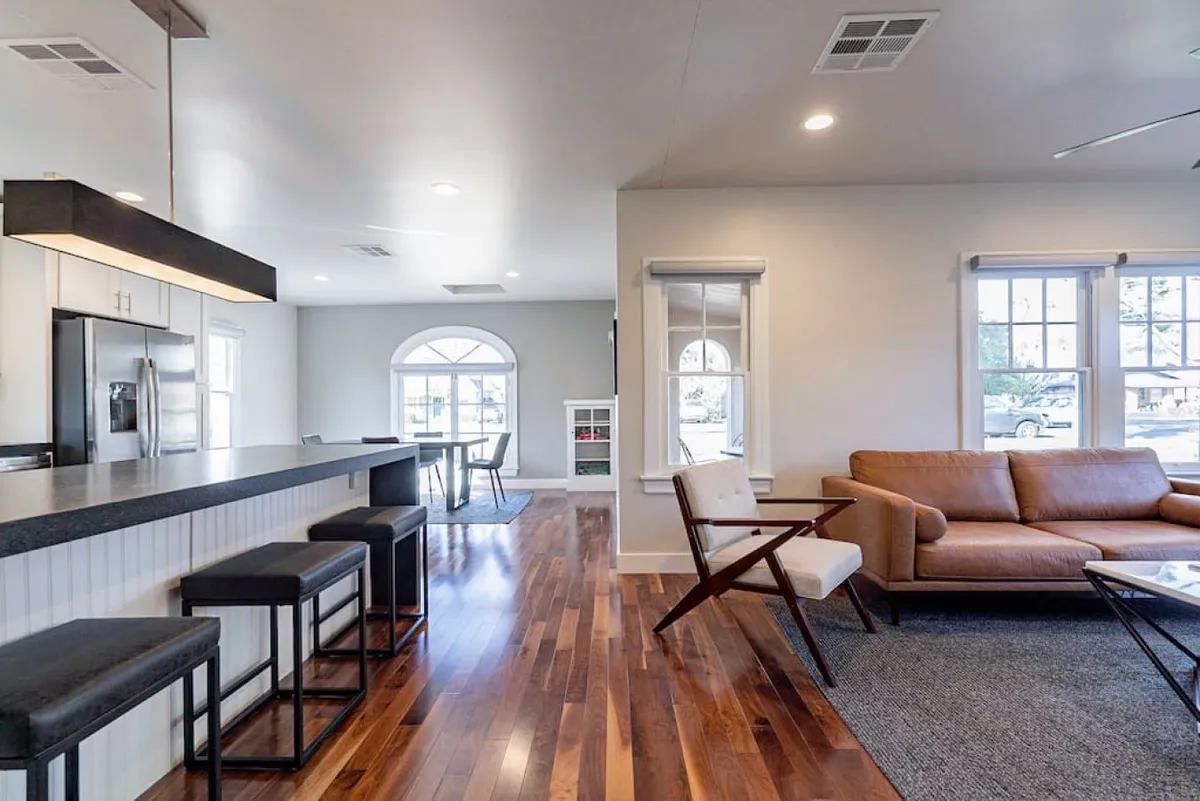 A bright, open-plan space featuring a kitchen island, comfortable sofa, and large windows. The scene exudes a welcoming and relaxed mood, with ample seating and sunlight creating an environment perfect for entertaining, reading, or family time.