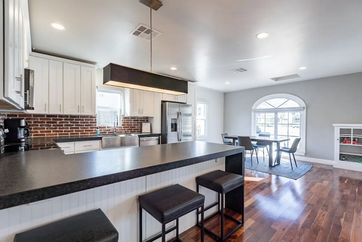 The image showcases a spacious, light-filled kitchen with a large island as the focal point, complemented by a stylish brick backsplash and a bright window that fills the space with natural light. Adjacent to the kitchen is a cozy dining area, inviting for meals and gatherings. The overall ambiance is both welcoming and functional, perfect for hosting family and friends, holiday celebrations, or casual dinners.