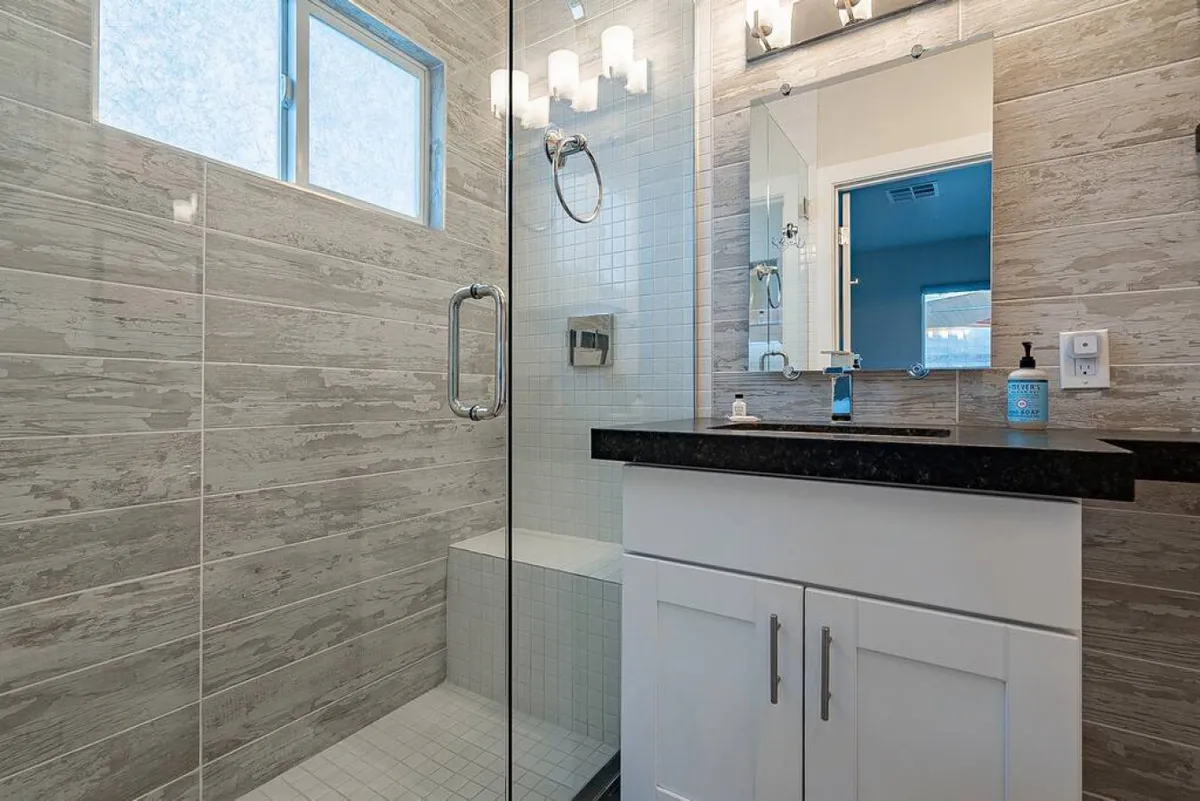 A freshly remodeled bathroom with a sleek glass shower enclosure, black countertop, bright lighting, and a wide mirror. The clean, modern fixtures and calming design suggest a spa-like atmosphere.