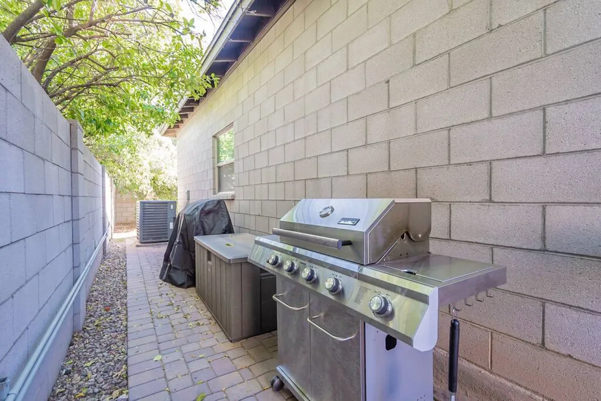 An outdoor grilling area with a built-in grill and counter, set along a pathway and surrounded by greenery. The scene evokes summer barbecues and group meals, with space for socializing and preparing food outdoors.